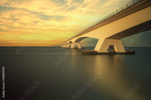 Zeeland Bridge - a long white bridge over the sea. The bridge is illuminated by beautiful evening light. Clouds illuminated by the sun. Dark surface of the river Oosterschelde.