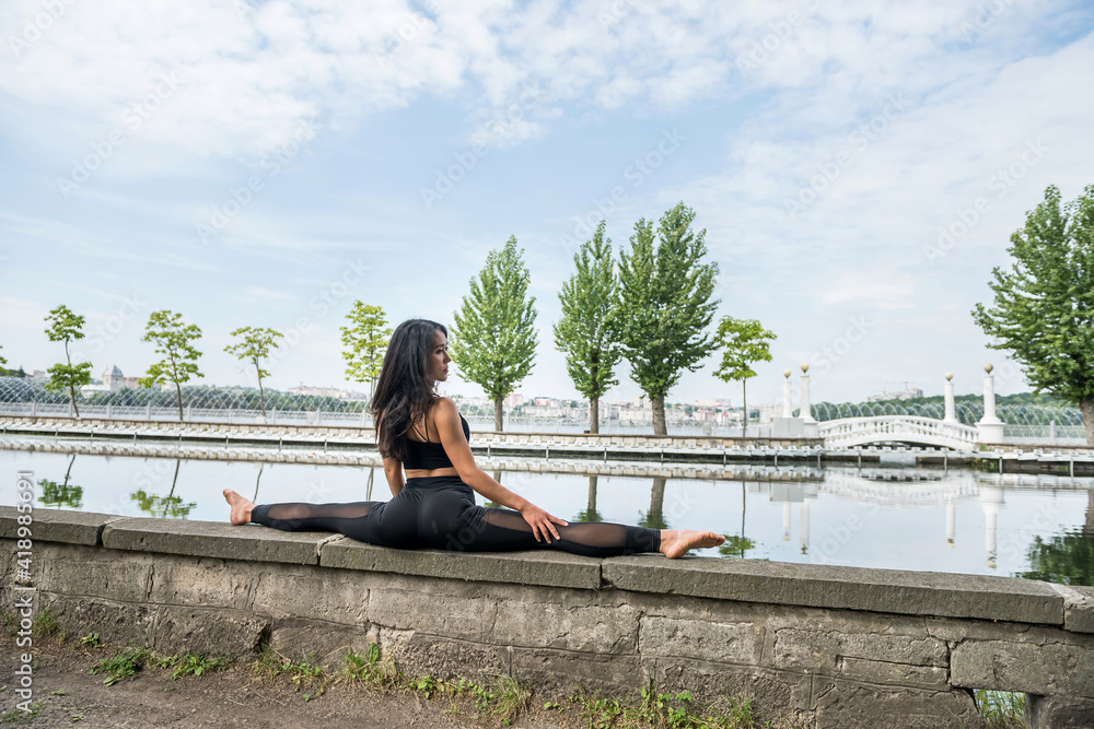 Young slim woman, wearing black sportswear sitting in twine and doing stretching exercises by lake Young slim woman, wearing black sportswear sitting in twine and doing stretching exercises by lake