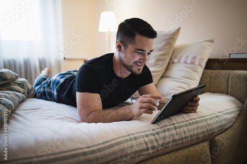 Young man having fun playing on tablet while laying in bed