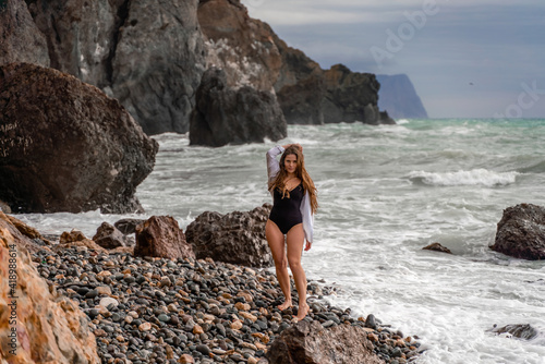 A beautiful girl in a white shirt and black swimsuit stands on the edge of a cliff, big waves with white foam. A cloudy stormy day at sea, with clouds and big waves hitting the rocks.