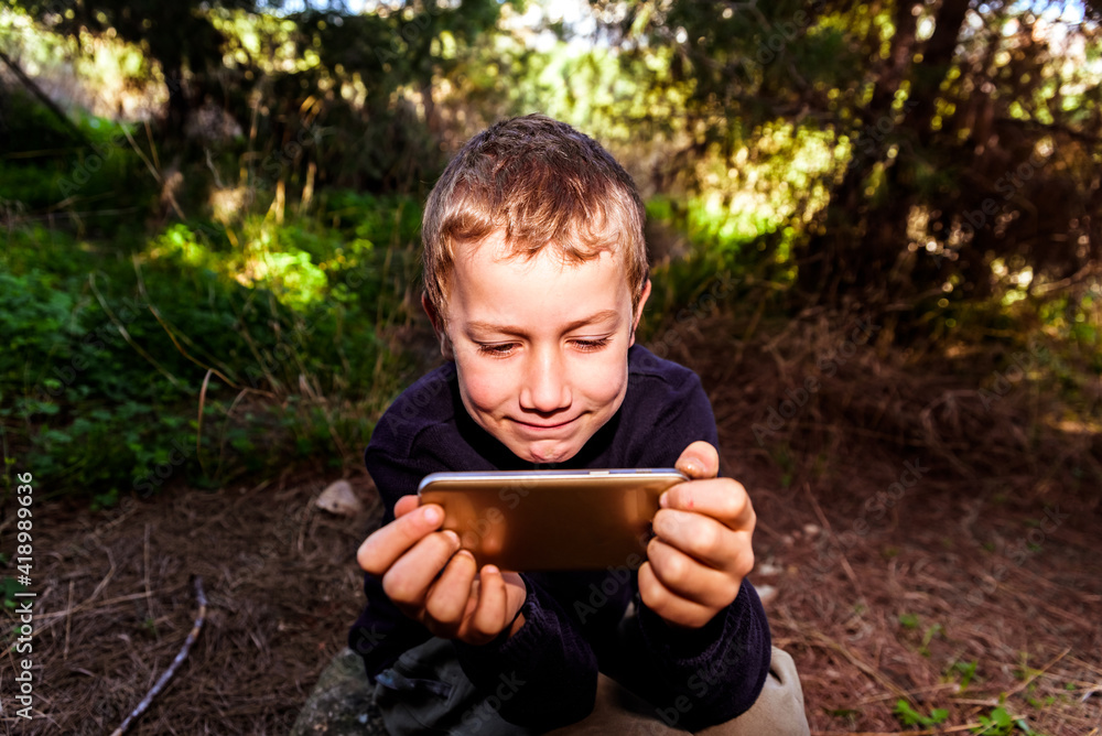A boy with an expression of great amusement has a great time watching videos on his smartphone while in a forest. A boy with an expression of great amusement has a great time watching videos on his smartphone while in a forest.