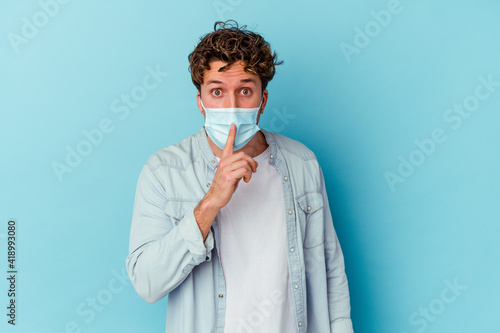 Young caucasian man wearing an antiviral mask isolated on blue background keeping a secret or asking for silence. © Asier