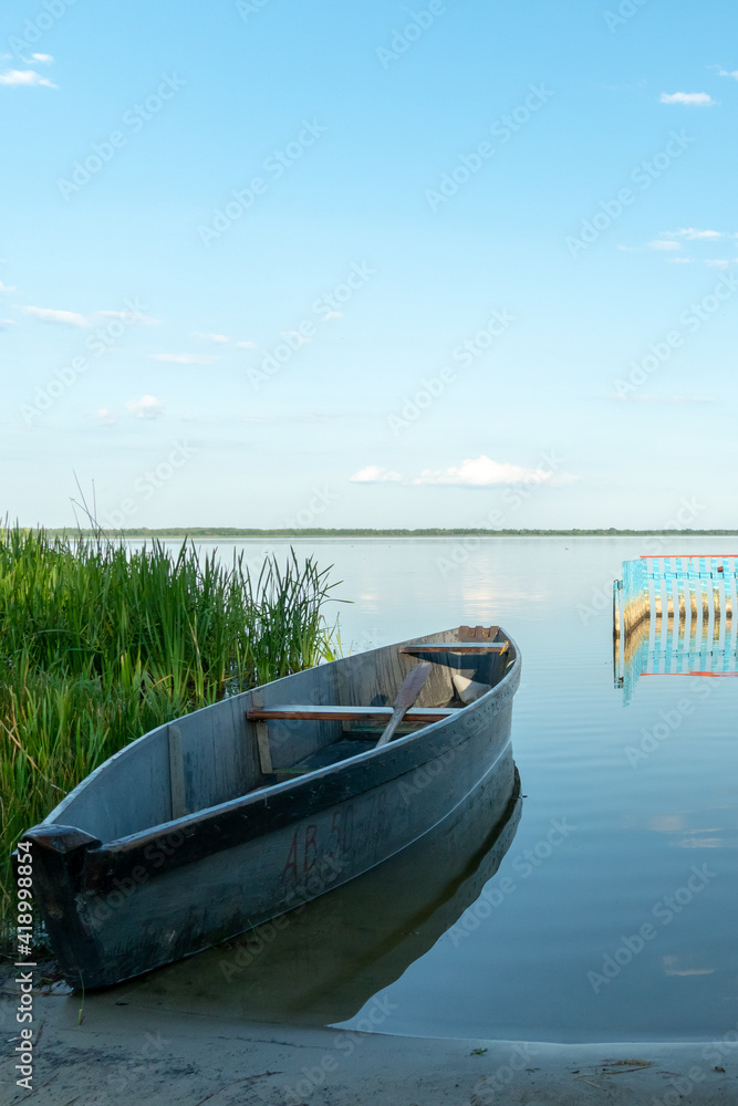 An old wooden fishing boat docked on a deserted sandy beach. View of a quiet and calm lake without ripples on the water. Reflection in the clear water of the blue sky and fluffy clouds during sunset. An old wooden fishing boat docked on a deserted sandy beach. View of a quiet and calm lake without ripples on the water. Reflection in the clear water of the blue sky and fluffy clouds during sunset.