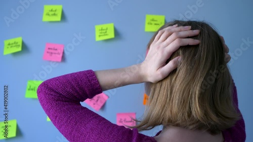 Blonde woman takes her head with her hands against the backdrop of large wall with stickers on which tasks and reminders are written. Concept: time management problems. photo