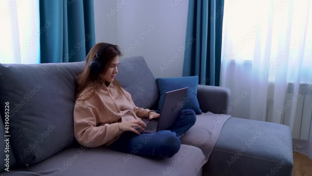 Smiling young girl in headphones using laptop while sitting on sofa at home. Happy girl looking at computer screen watching webinar, online language learning, distance education concept. Smiling young girl in headphones using laptop while sitting on sofa at home. Happy girl looking at computer screen watching webinar, online language learning, distance education concept.