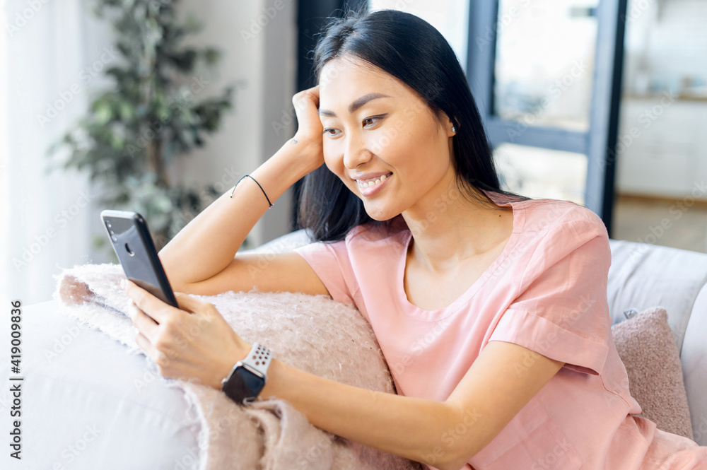 Asian woman mixed-race pretty lady sitting on the couch, looking at mobile phone, video-calling, texting, talking to friend, family, taking selfie for social media, staying connected during pandemic Asian woman mixed-race pretty lady sitting on the couch, looking at mobile phone, video-calling, texting, talking to friend, family, taking selfie for social media, staying connected during pandemic