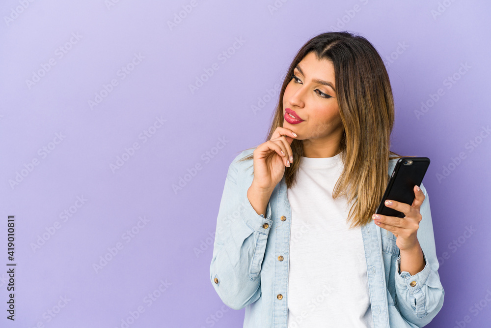 Young indian woman holding a phone isolated looking sideways with doubtful and skeptical expression. Young indian woman holding a phone isolated looking sideways with doubtful and skeptical expression.