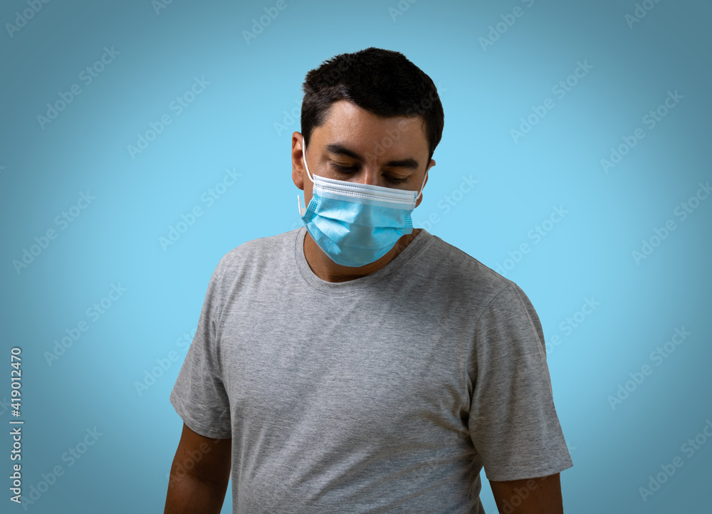Brunette man wearing a covid pandemic protective mask with his hands on his waist, wearing a gray t-shirt, looking down on a light blue background. Expression of tired and sad. Brunette man wearing a covid pandemic protective mask with his hands on his waist, wearing a gray t-shirt, looking down on a light blue background. Expression of tired and sad.