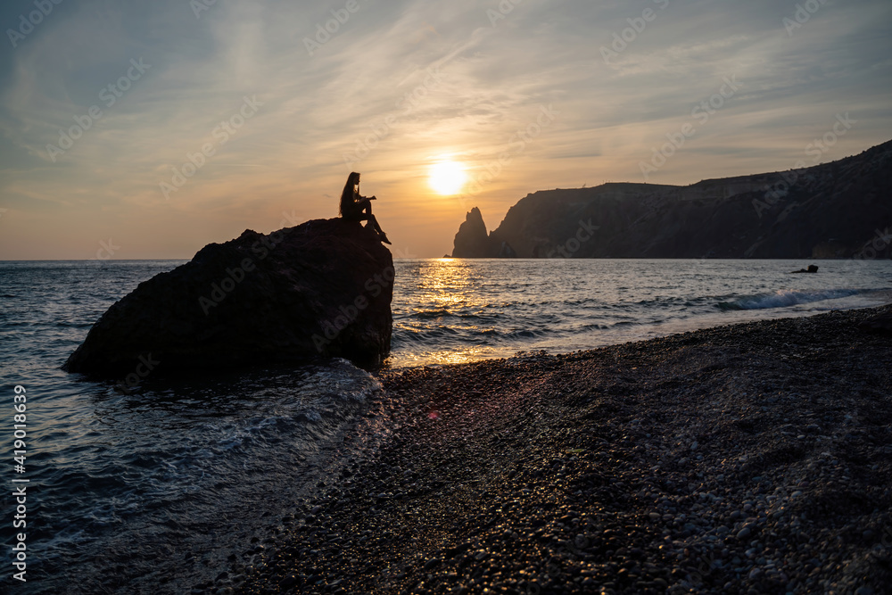 A girl on the beach watches the sunset on the sea. She's sitting on a rock in the sea. The sun is setting behind the mountains.
