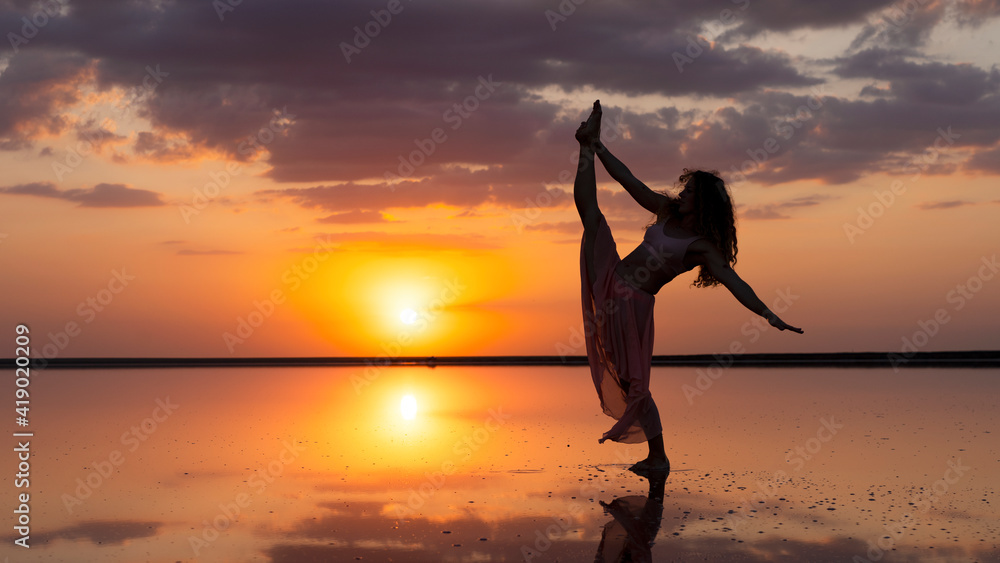 Beautiful girl among clouds in airy dress performs split on background of incredible sunset sky. silhouette of dancer, mirror surface of salt lake