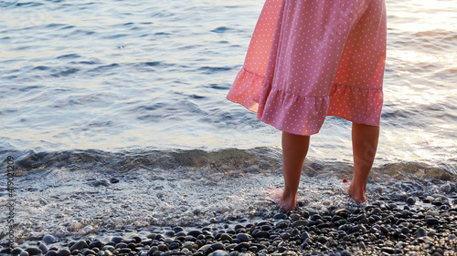 Woman's legs in pink dress stand barefoot in water on sea pebble shore