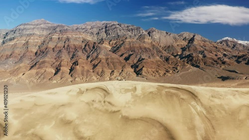 People hiking to the top of high sand dune with painted mountains landscape on motion background. Tourists walking by golden dune hills under blue sky. Nature 4K. Travelers exploring wilderness desert