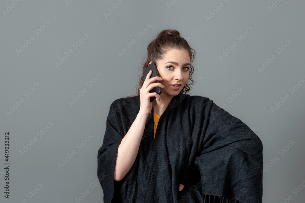 curly-haired emotional young woman in a black cloak talk on mobile phone, isolated on gray curly-haired emotional young woman in a black cloak talk on mobile phone, isolated on gray