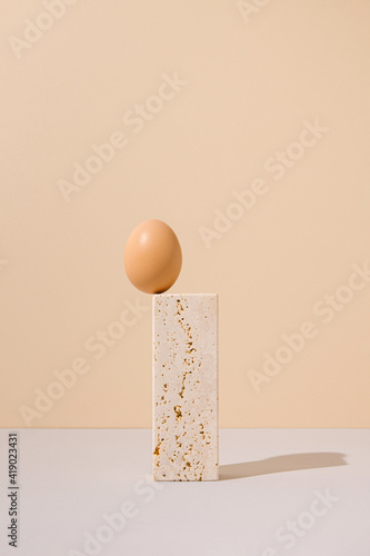 Single egg standing on a travertine marble block on a beige and gray background. 2021 Easter unique still life arrangement. Surreal retro creative concept.