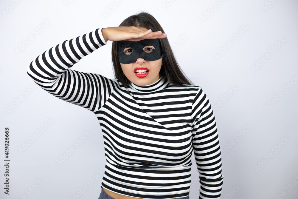 Young beautiful brunette burglar woman wearing mask very happy and smiling looking far away with hand over head. searching concept. Young beautiful brunette burglar woman wearing mask very happy and smiling looking far away with hand over head. searching concept.