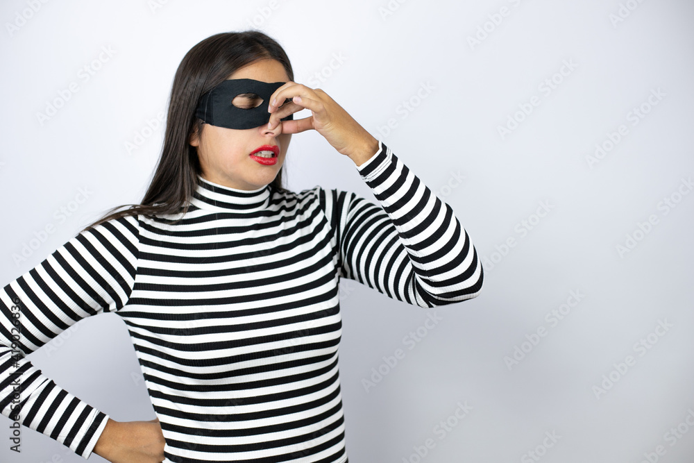 Young beautiful brunette burglar woman wearing mask smelling something stinky and disgusting, intolerable smell, holding breath with fingers on nose Young beautiful brunette burglar woman wearing mask smelling something stinky and disgusting, intolerable smell, holding breath with fingers on nose
