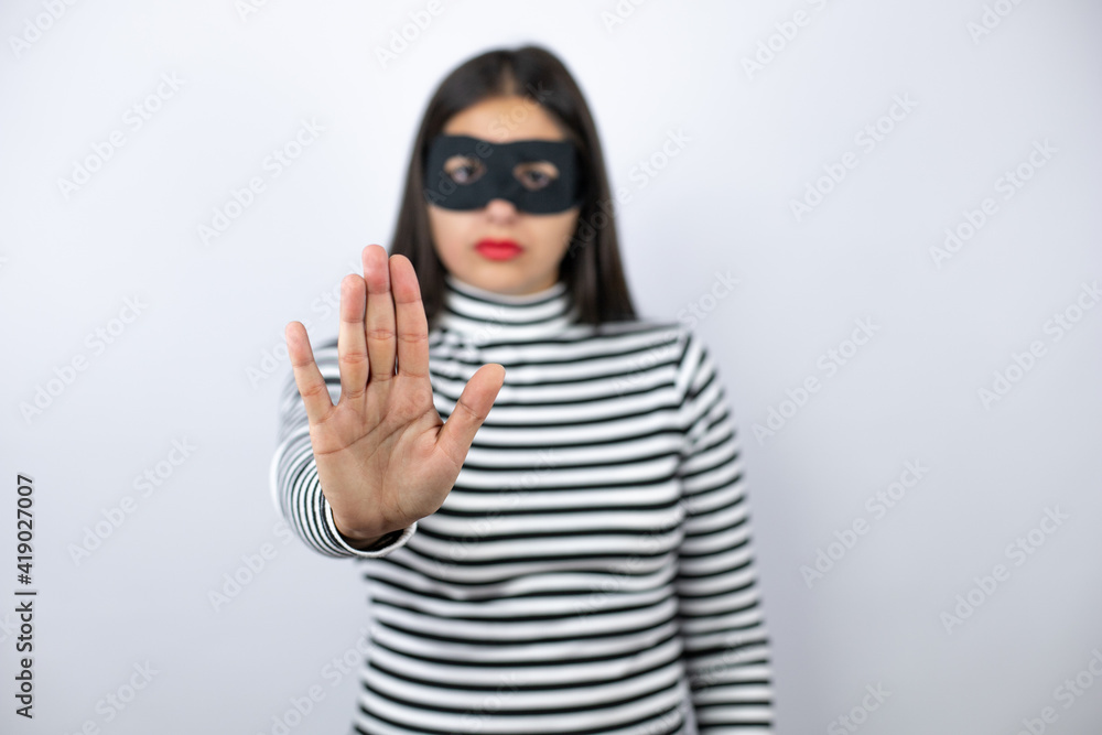 Young beautiful brunette burglar woman wearing mask serious and doing stop sing with palm of the hand. Young beautiful brunette burglar woman wearing mask serious and doing stop sing with palm of the hand.