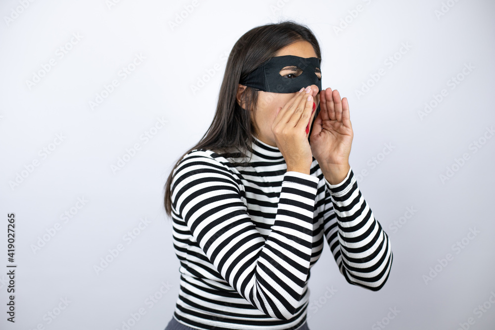 Young beautiful brunette burglar woman wearing mask shouting and screaming loud to side with hand on mouth Young beautiful brunette burglar woman wearing mask shouting and screaming loud to side with hand on mouth