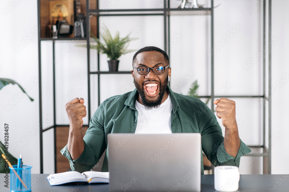 Happy African American male freelancer dressed in stylish clothes, sitting at his work desk using laptop, shouts and rejoice in success, win or good profit, looking at the camera and gesturing hands