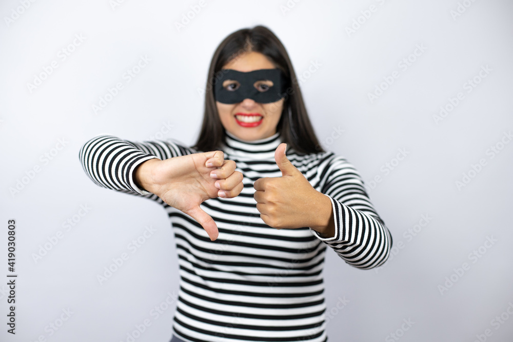 Young beautiful brunette burglar woman wearing mask Doing thumbs up and down, disagreement and agreement expression. Crazy conflict Young beautiful brunette burglar woman wearing mask Doing thumbs up and down, disagreement and agreement expression. Crazy conflict