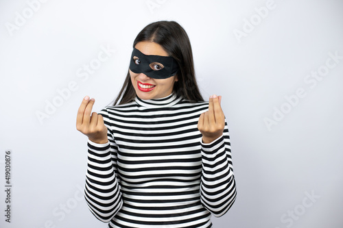 Young beautiful brunette burglar woman wearing mask doing money gesture with hands, asking for salary payment, millionaire business