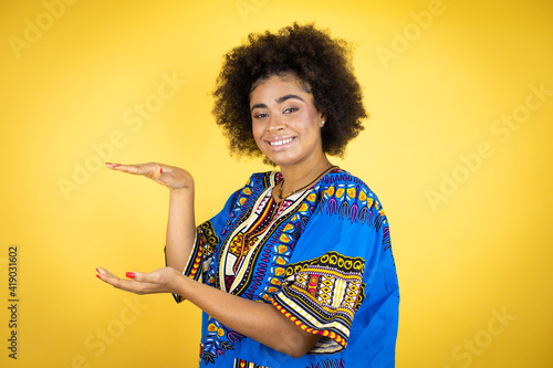 African american woman wearing african clothing over yellow background gesturing with hands showing big and large size sign, measure symbol. Smiling . Measuring concept.