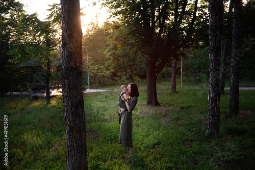 Young mother holds baby in arms in the rays of the sunset in the park between pine trees green grass. Woman kisses, hugs girl daughter. Maternal care, custody. Adoption concept. Walks in the open air