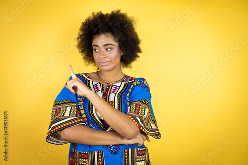 African american woman wearing african clothing over yellow background confused and pointing with hand and finger to the side