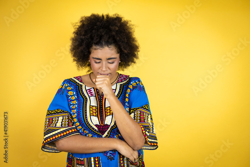 African american woman wearing african clothing over yellow background with her hand to her mouth because she's coughing