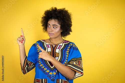 African american woman wearing african clothing over yellow background surprised and pointing her fingers side
