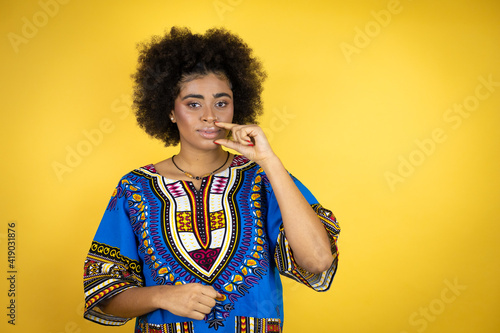 African american woman wearing african clothing over yellow background mouth and lips shut as zip with fingers. Secret and silent, taboo talking