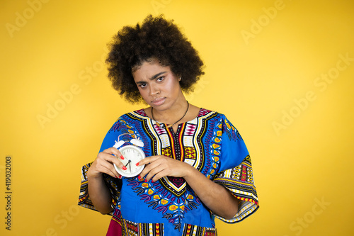 African american woman wearing african clothing over yellow background serious and holding and pointing a clock