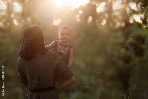 Young mother holds baby in arms in the rays of the sunset in the park, the girl looks out from behind. The woman kisses, hugs daughter. Maternal care, custody. Adoption concept. Walks in the open air