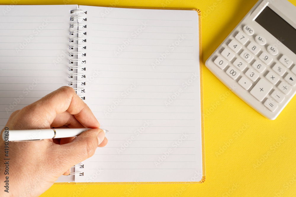 close up on hand holding pencil writing on notebook by the calculator on yellow background - office and business or goals concept close up on hand holding pencil writing on notebook by the calculator on yellow background - office and business or goals concept
