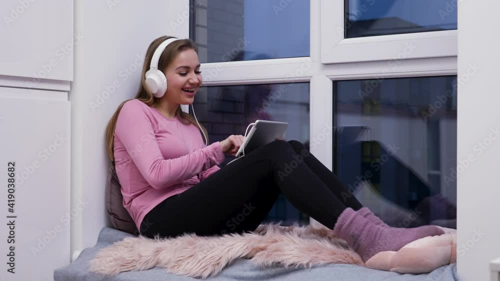 Young woman sits on windowsill near large window wearing big white headphones, watching movie on tablet, rejoices and laughs. Female in home clothes poses against backdrop of light room. Slow motion. Young woman sits on windowsill near large window wearing big white headphones, watching movie on tablet, rejoices and laughs. Female in home clothes poses against backdrop of light room. Slow motion.