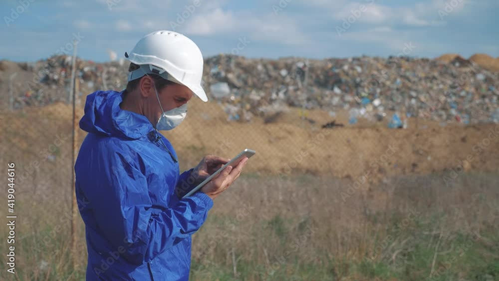 environmental pollution. man chemist scientist in a respirator mask studies landfill dump pollution digital tablet and an white helmet stands next to standing near the mountains of garbage. lifestyle environmental pollution. man chemist scientist in a respirator mask studies landfill dump pollution digital tablet and an white helmet stands next to standing near the mountains of garbage. lifestyle