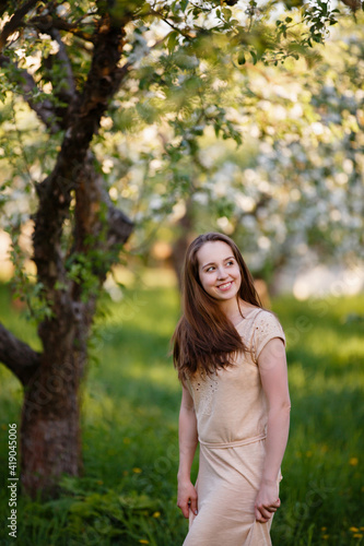 Young woman in beige dress in a blooming apple garden. Spring story. Brown-haired girl with long hairs. Woman plays with her hairs