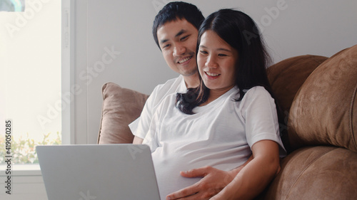 Young Asian Pregnant couple using laptop search pregnancy information. Mom and Dad feeling happy smiling positive and peaceful while take care their child lying on sofa in living room at home concept.