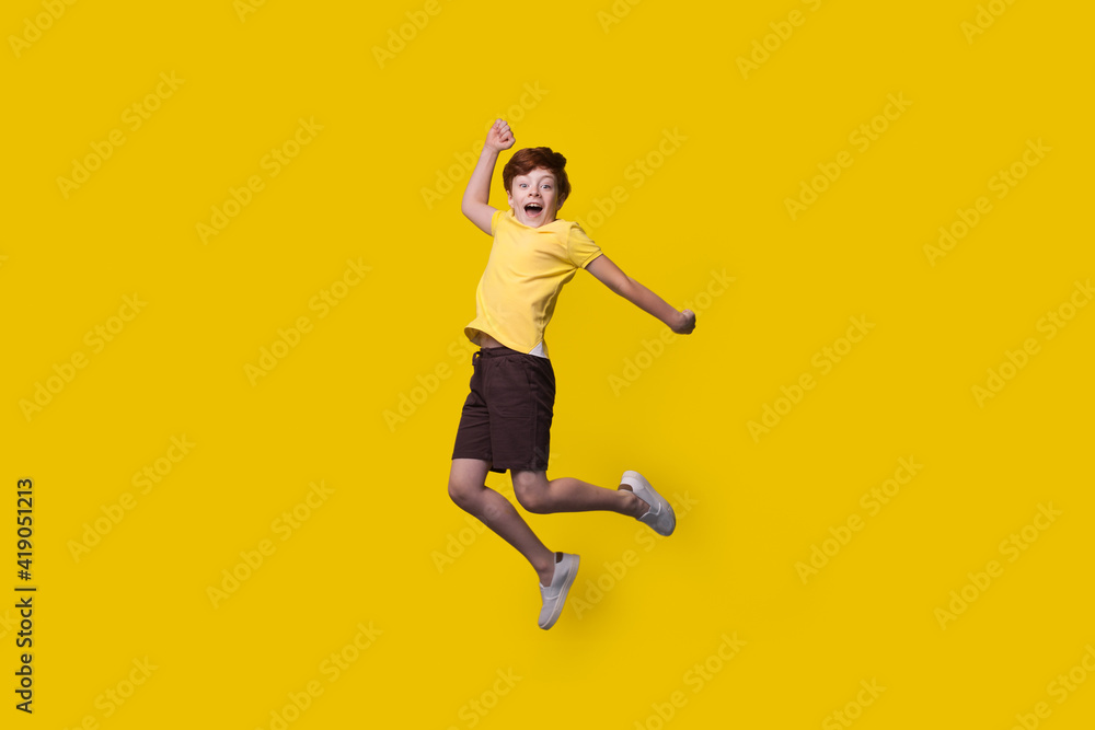 Ginger caucasian boy is jumping on a yellow studio wall with free space smiling at camera