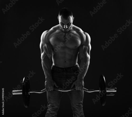 Muscular strong men, athlete, bodybuilder, weightlifter does exercises for for top and arms, working out with barbell in gym over dark background. Young man lifting weights. Black and white 