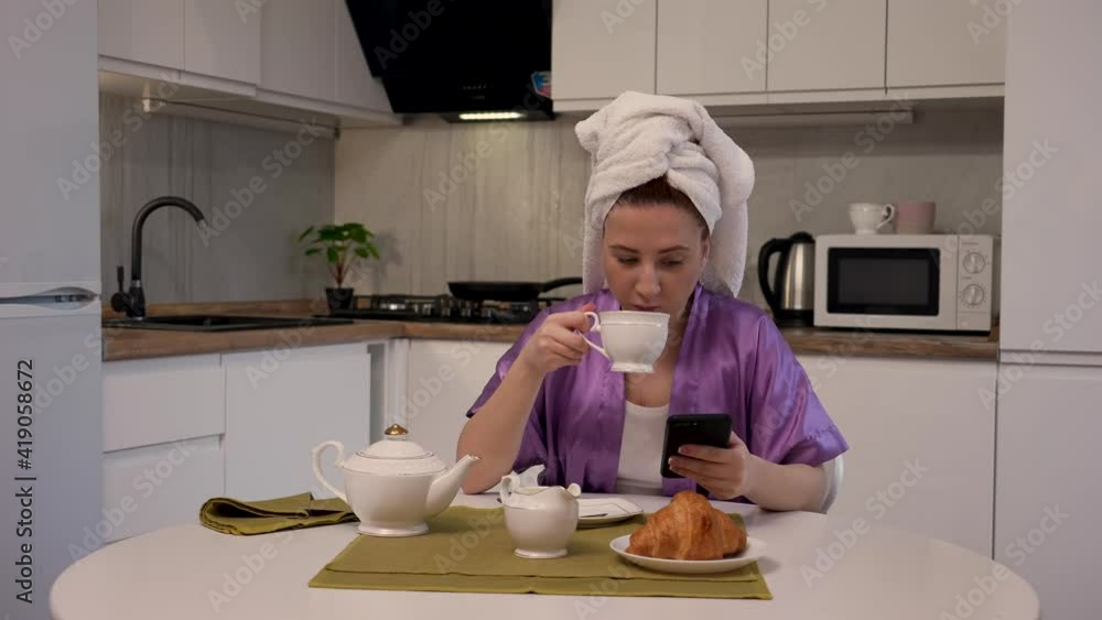 Woman is sitting at table in kitchen, makes an online purchase using smartphone and credit card, drinks coffee. Female in white towel on her head poses against background of light room. Slow motion. Woman is sitting at table in kitchen, makes an online purchase using smartphone and credit card, drinks coffee. Female in white towel on her head poses against background of light room. Slow motion.