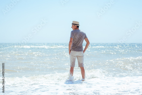 Rear view of young man in a gray shirt, white shorts and straw hat standing on empty beach at sunny midday. A male stands in the waves of the sea and looking aside. Alone on a beach admiring the waves