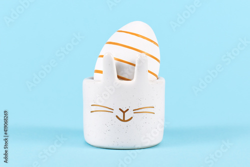 White Easter egg painted with golden stripes in cute bunny shaped egg cup on blue background