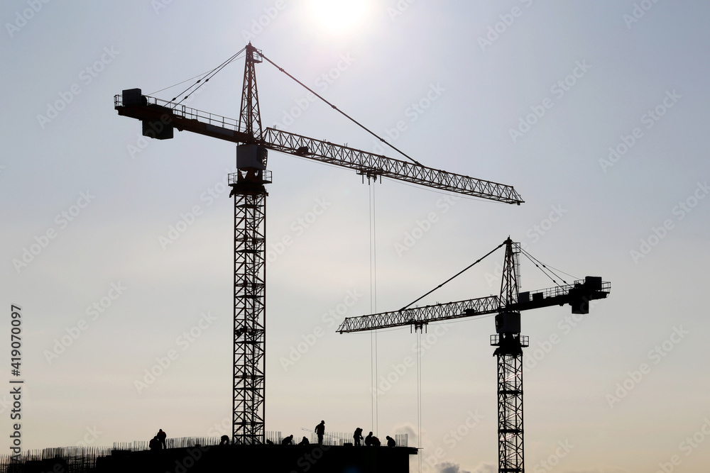 Silhouettes of construction cranes and workers on unfinished residential building against sky and sunshine. Housing construction, apartment block in city Silhouettes of construction cranes and workers on unfinished residential building against sky and sunshine. Housing construction, apartment block in city
