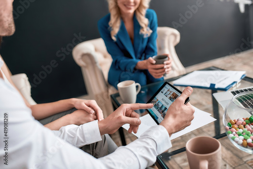 Close up shot of male client holding tablet pc while choosing tour, sitting with his girlfriend in modern travel agency office