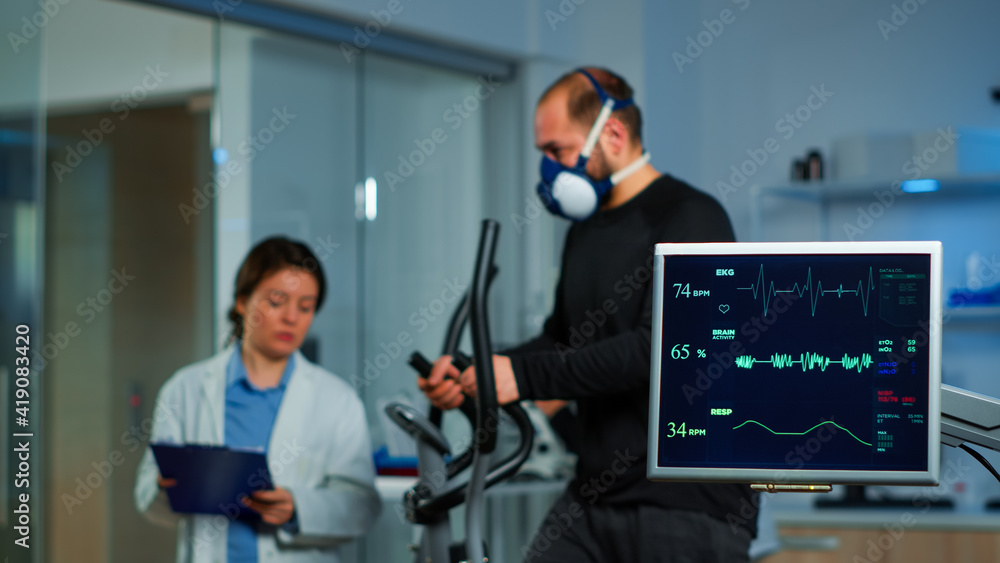 Team of medial researchers monitoring vo2 of man performance sports wearing mask running. Lab science doctor measuring endurance of sportsman while ekg scan runs on computer screen in laboratory Team of medial researchers monitoring vo2 of man performance sports wearing mask running. Lab science doctor measuring endurance of sportsman while ekg scan runs on computer screen in laboratory
