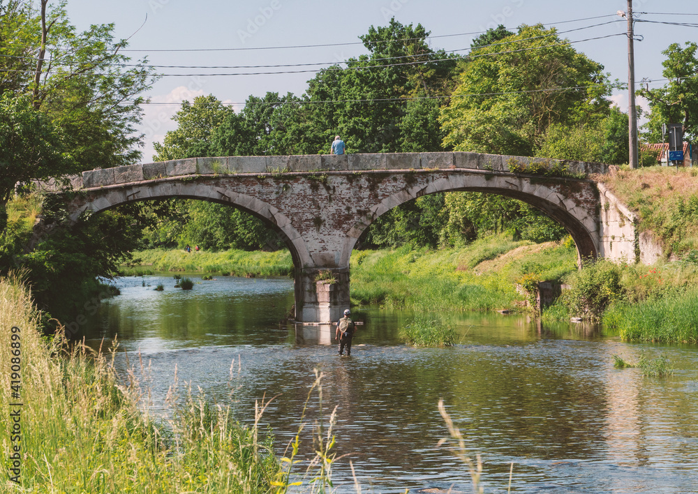 Fototapeta premium picturesque stretch of the Ticino river with ancient stone bridge frequented by fishermen in the summer season. Ticino Park, Lombardy, Italy.