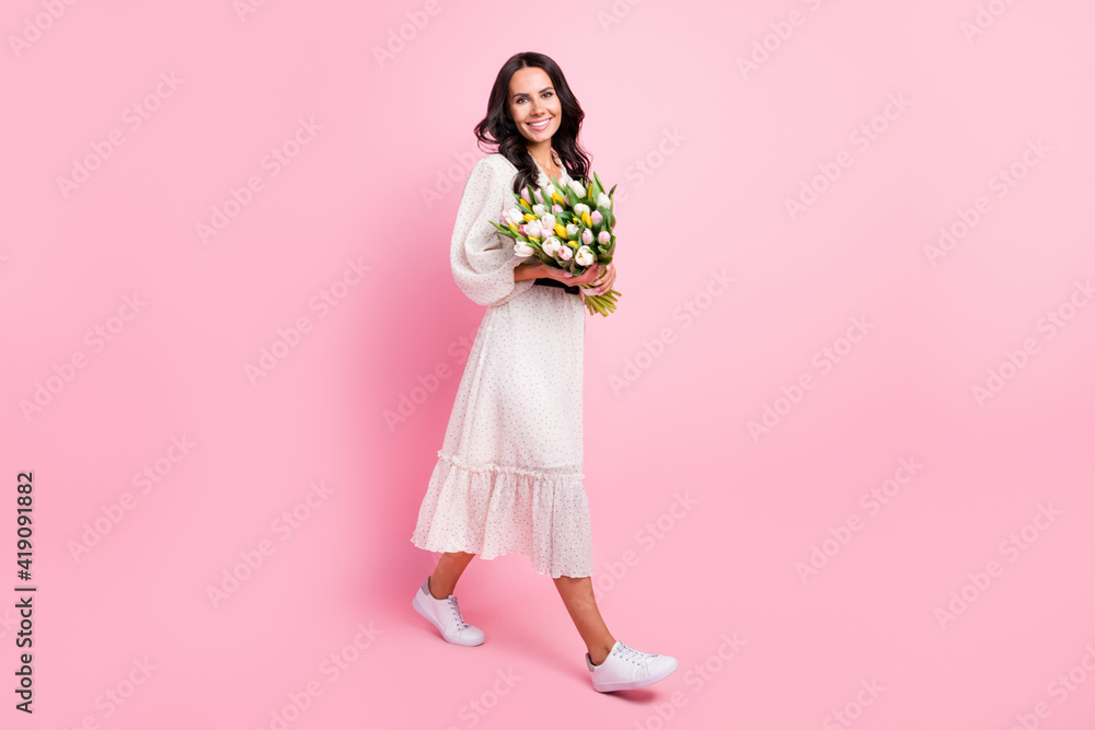 Full body profile side photo of charming nice woman walk empty space smile hold flowers spring isolated on pink color background Full body profile side photo of charming nice woman walk empty space smile hold flowers spring isolated on pink color background