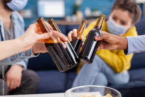 Close up shot of mixed race friends clinking beer bottles hanging out keeping social distancing having fun in apartment linving room during global pandemic. Conceptual image. photo