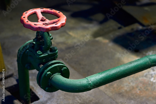 Industrial green chemical petrochemical retro style valve closeup with selective focus on red steering wheel mounted with flange connection on green pipe, out of focus foreground with copyspace.